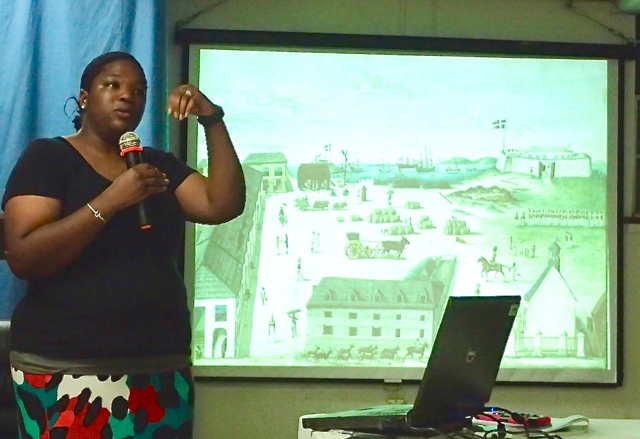 Doctoral candidate Alicia Odewale talks about the Slave Wrecks Project excavation with an artist rendering of the Christiansted National Historic Site warehouse compound during the 1700&rsquo;s in the background.